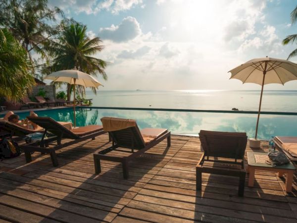 A serene poolside scene with lounge chairs, umbrellas, palm trees, and an ocean view under a bright sky.