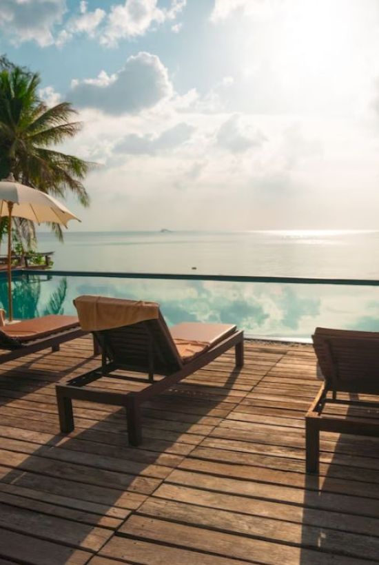 A serene poolside scene features lounge chairs and umbrellas with a picturesque view of the ocean and palm trees under a bright sky.