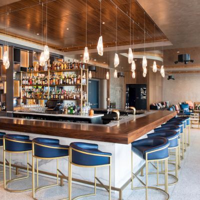The image shows a modern bar with a central countertop, surrounded by blue stools and warm lighting, creating a cozy atmosphere.