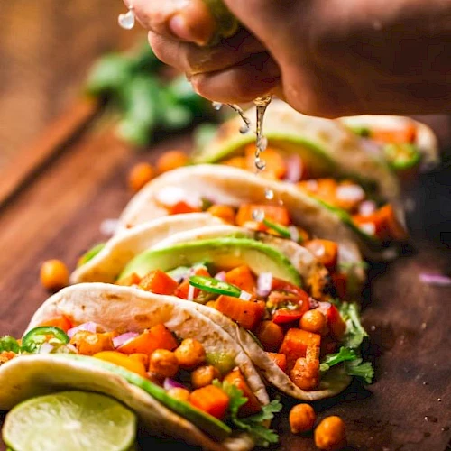 A hand squeezes lime over tacos filled with chickpeas, avocado, and veggies on a wooden board, with lime slices in the foreground.