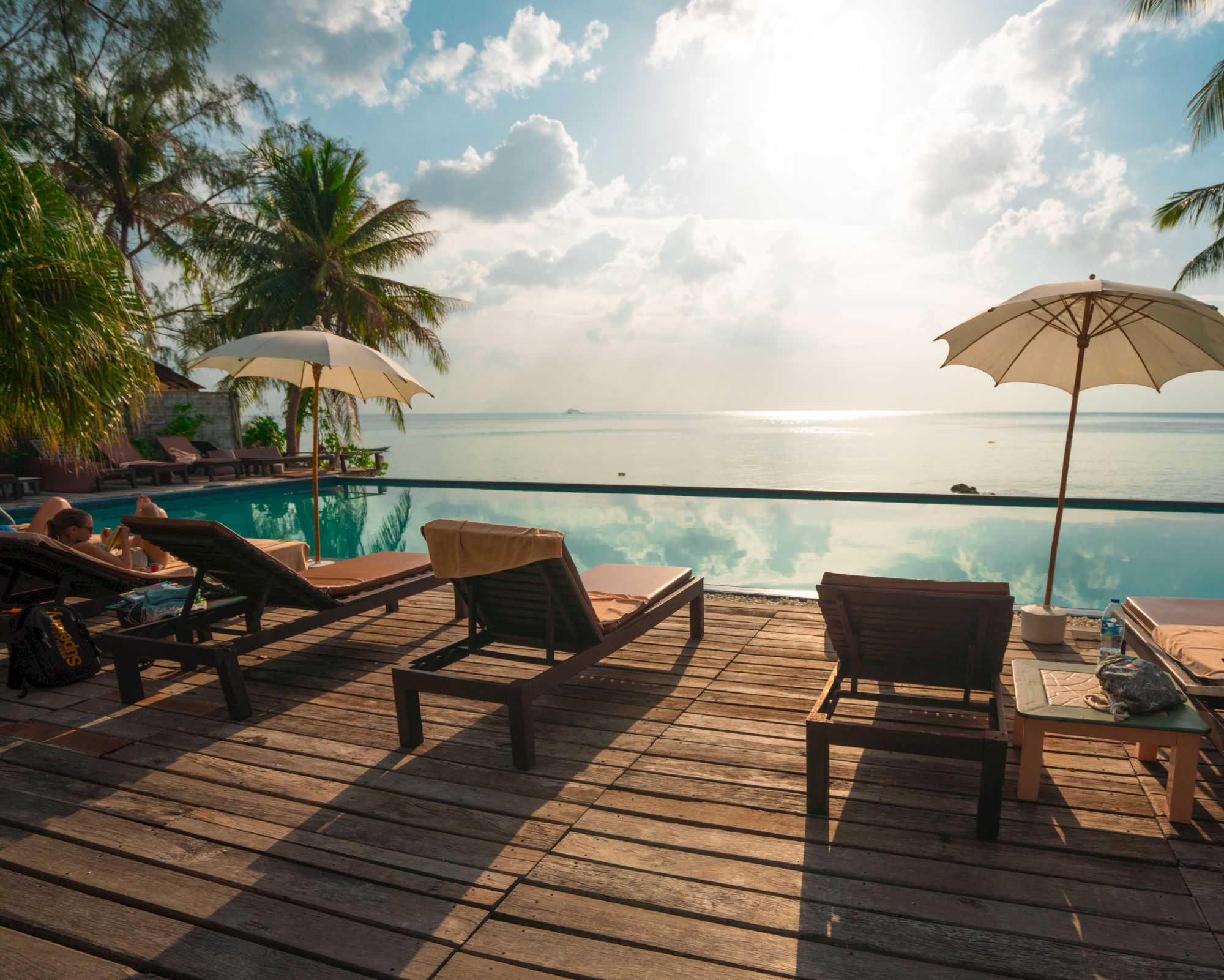 The image shows a beach setting with lounge chairs, umbrellas, and a tranquil sea view under a partly cloudy sky.
