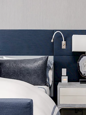 A modern bedroom features a bed with blue accents, a nightstand with a lamp and a phone, and a light gray wall in the background.