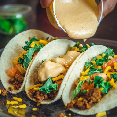Three tacos filled with ground meat, lettuce, cheese, and tomatoes are being topped with a creamy sauce on a plate.