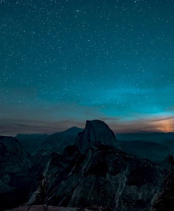 A starry night sky over a mountainous landscape with silhouettes of trees and a prominent peak in the foreground.