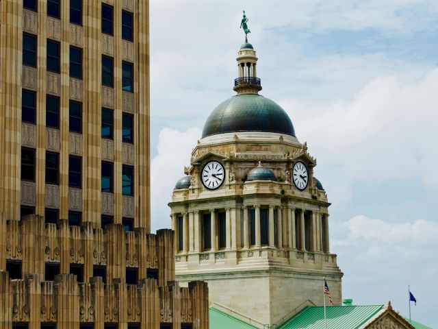 A cityscape featuring a historic cathedral with Gothic spires and a domed, green-roofed building beneath a partly cloudy sky.