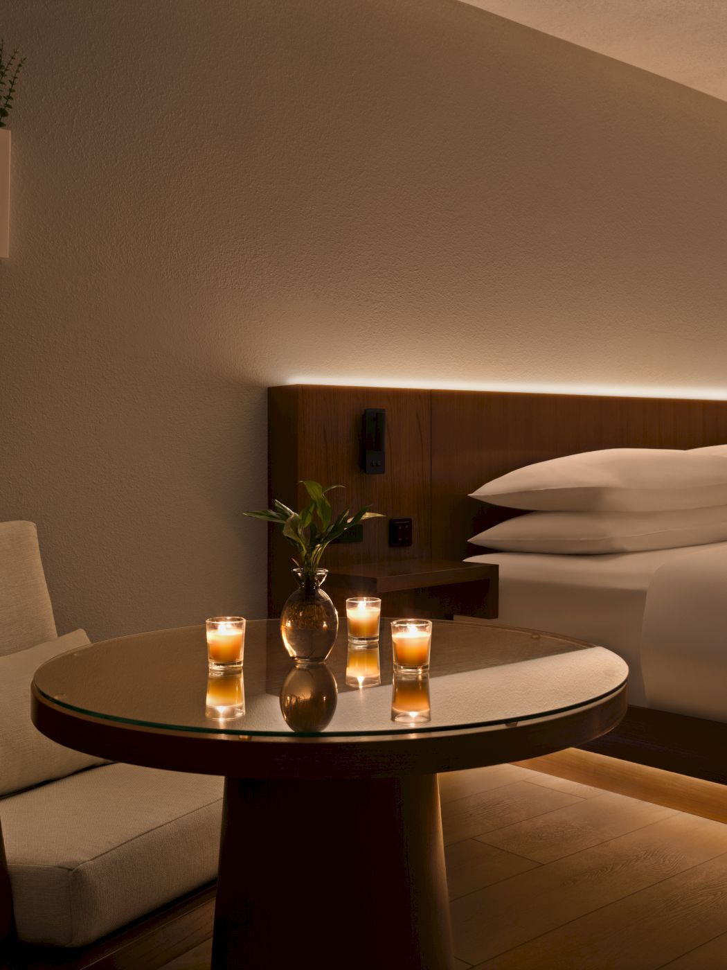 A cozy hotel room with a round table, two chairs, lit candles, and a neatly made bed against a warm, softly lit wall.
