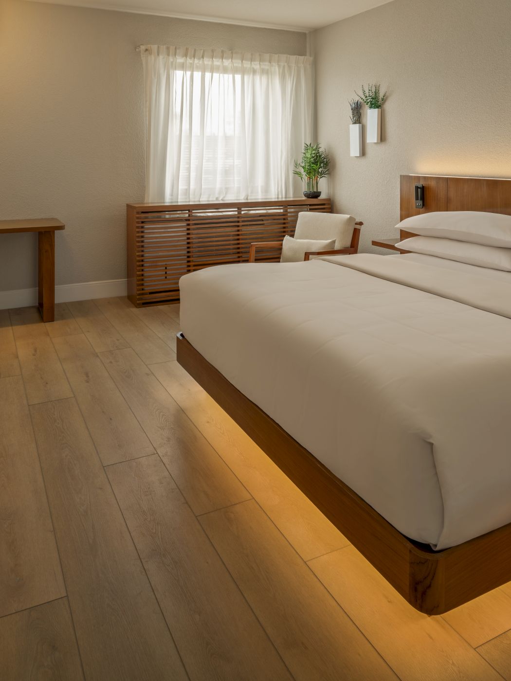 A modern hotel room with a large bed, wooden furniture, soft lighting, a desk, chair, plant, and warm under-bed lighting.