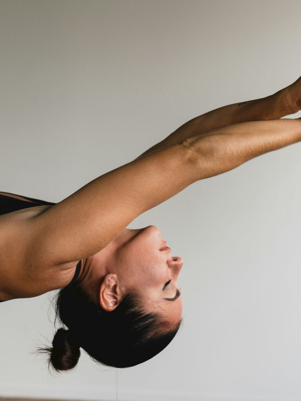 A person doing a forward bending yoga pose with arms extended, wearing a dark sports top, against a plain background.