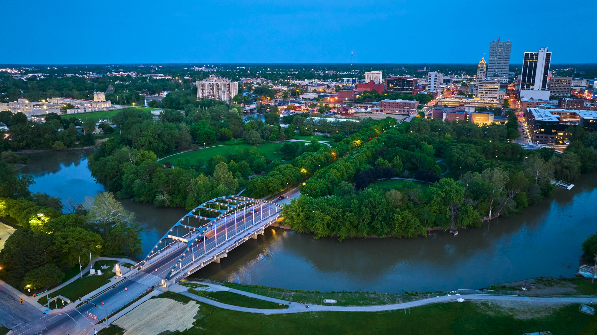 Aerial view of a city with a curved river, lush green park, a white arched bridge, and surrounding buildings in the distance, under a blue sky.