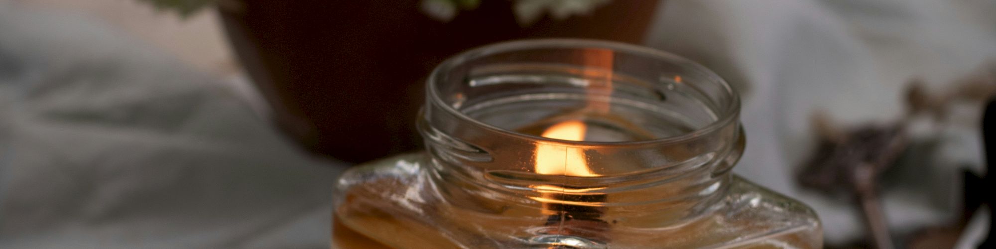 A lit candle in a glass jar sits on a table with an envelope and flowers in a pot in the background.