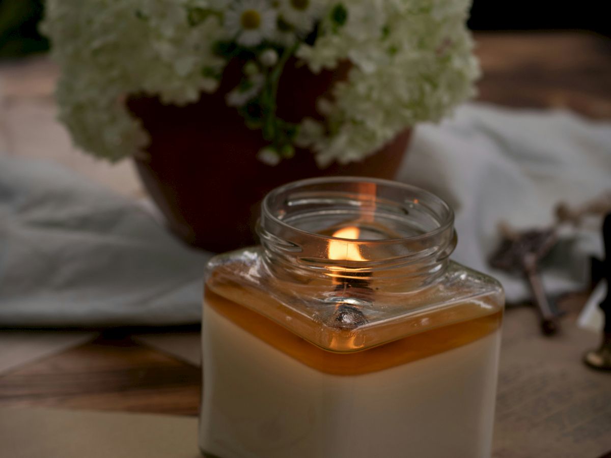 A lit candle in a glass jar sits on a wooden table next to an envelope and a potted plant with white flowers.