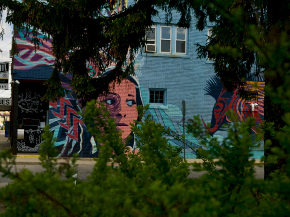 A colorful mural is painted on a building wall, featuring a face and abstract designs, partially obscured by trees in the foreground.