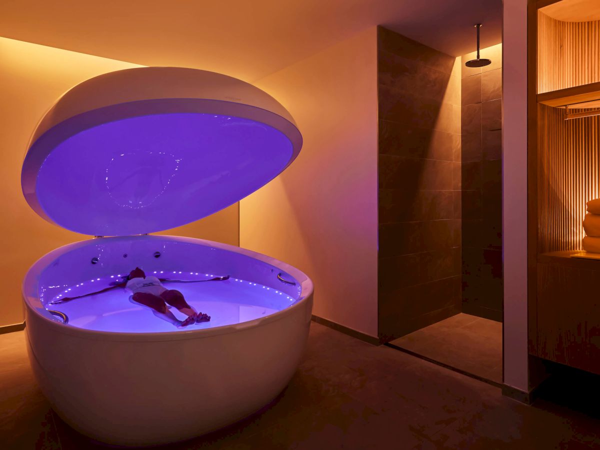 A person is floating in an open sensory deprivation tank with blue lighting, in a room with warm lighting and a shower area nearby.