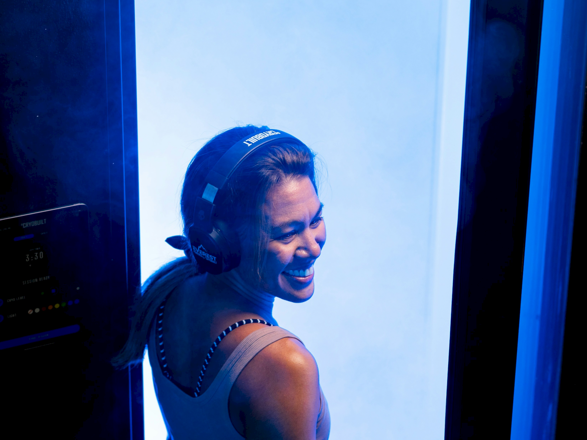 A person is smiling while stepping out of a cryotherapy chamber with a blue glow, wearing headphones and gloves.