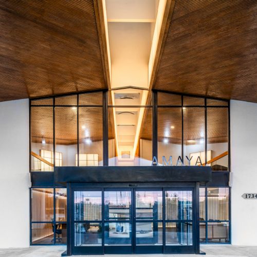 Modern exterior arrival at The Amaya hotel in Fort Wayne, featuring a glass fa&ccedil;ade and warm interior lighting at dusk.