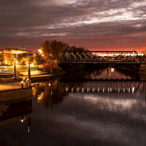 Promenade Park in Fort Wayne at dusk with illuminated riverwalk and bridge reflections
