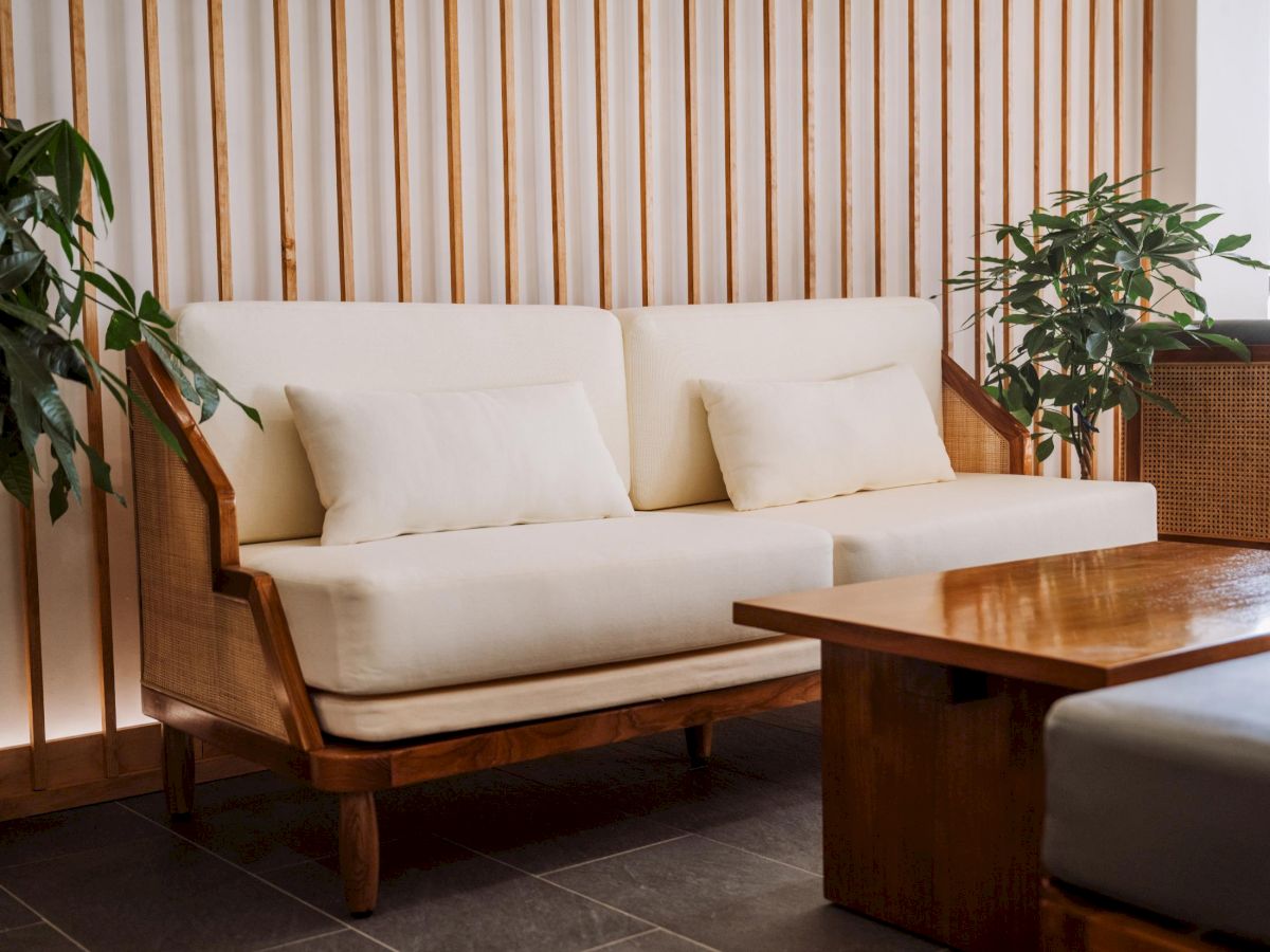 A cozy living room with a cream sofa, wooden armchairs, a wooden coffee table, striped wall backdrop, and potted plants on either side.