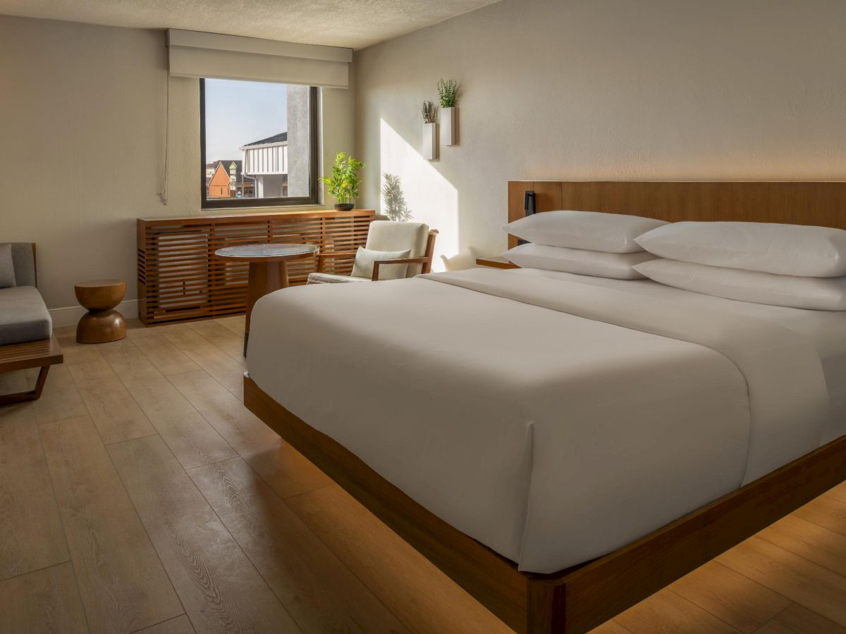 A modern hotel room with a large wooden bed, white linens, a seating area by the window, and warm ambient lighting.