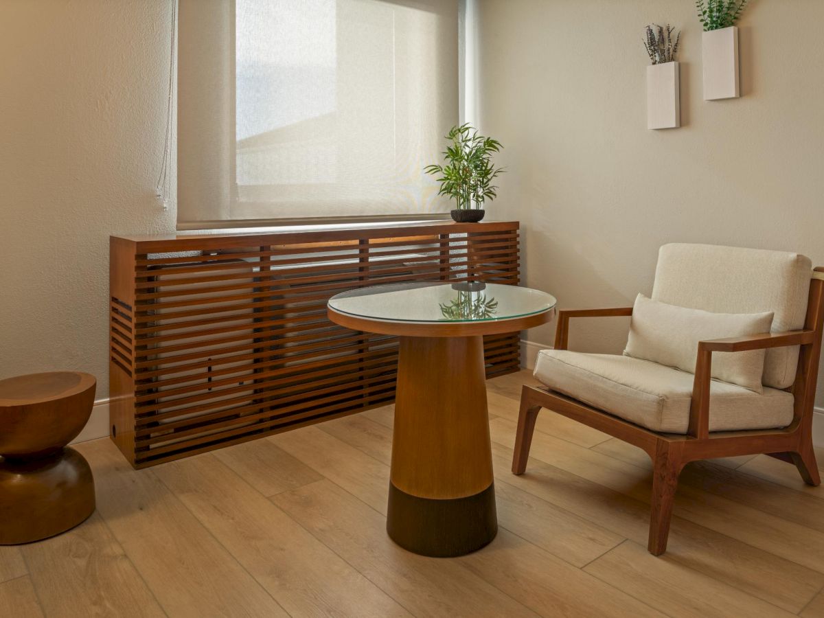 A cozy, minimal living room with a wooden chair, round glass-top table, cylindrical wood base, a wooden cabinet by the window, a small stool, and potted plants along the wall, all in warm natural tones.