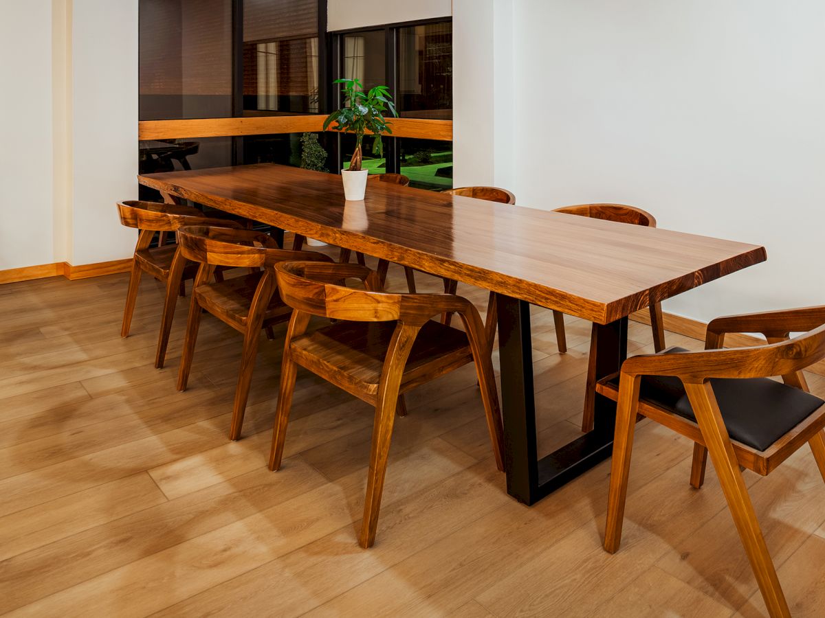 A long wooden dining table with eight matching chairs in a modern, minimal room; a small plant centerpiece and potted plant in the corner.