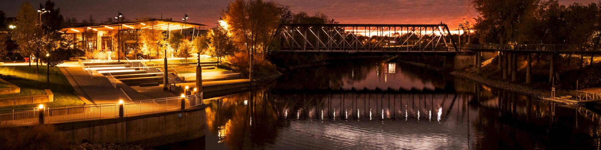 A calm river at dusk with a lit park and bridge, glowing reflections on the water, and a dark cloudy sky in the background.