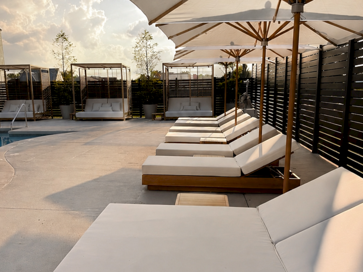 A modern outdoor poolside patio with loungers, umbrellas, privacy slats, and a backyard garden view under sunny skies.