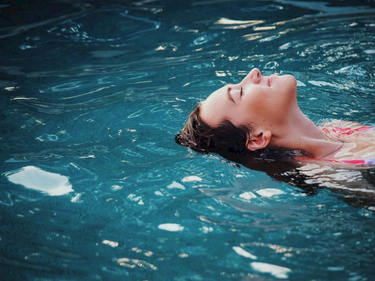 A person floats face-down in clear blue water, relaxing like a swimmer, with ripples sparkling in the pool.