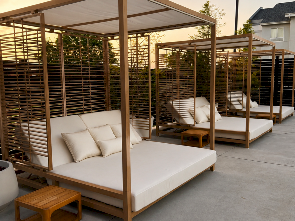 A modern outdoor patio with cushioned wooden daybeds under a pergola, plus soft evening lighting and a nearby house.