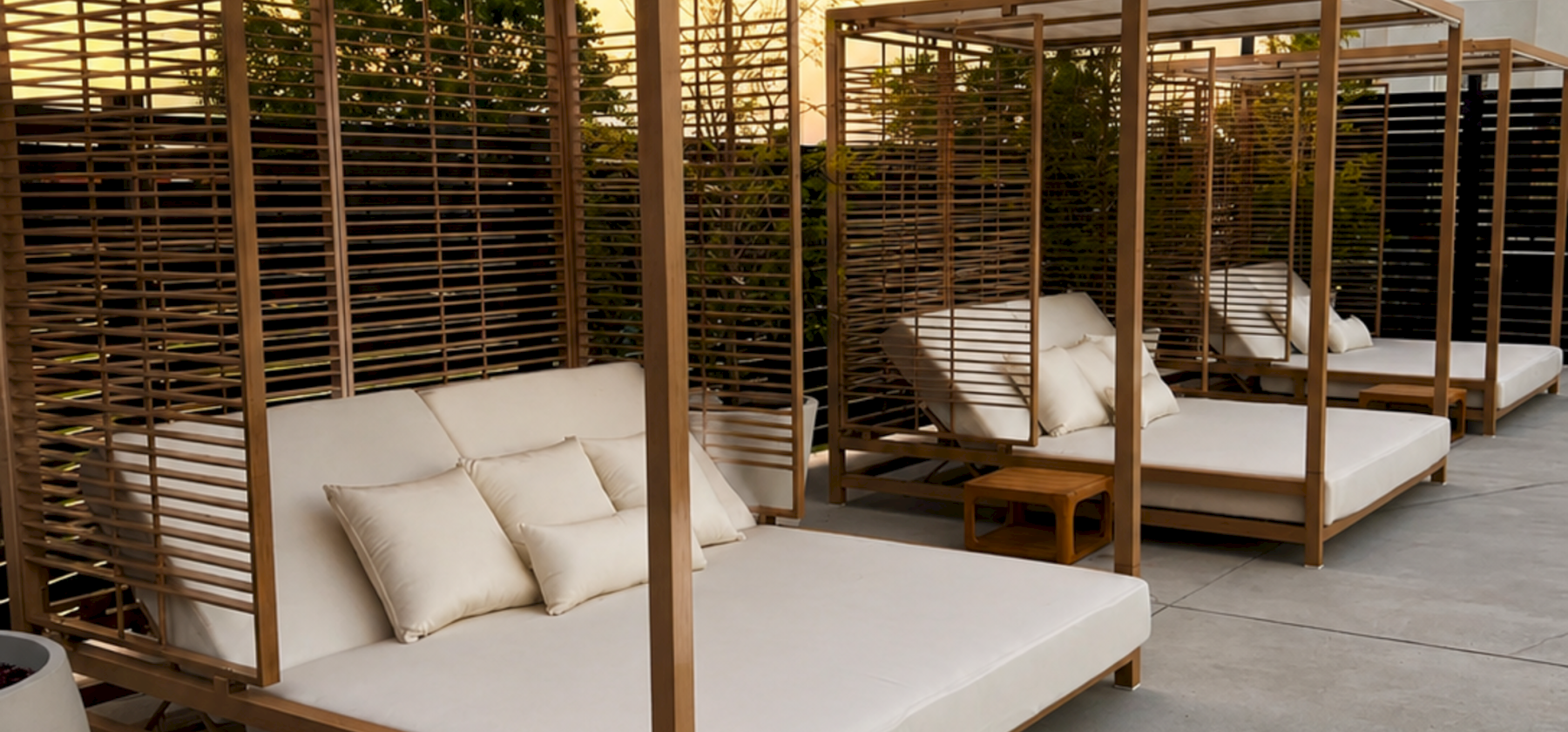 A modern outdoor patio with cushioned wooden daybeds under a pergola, plus soft evening lighting and a nearby house.