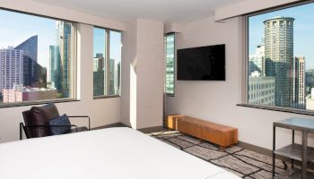 A modern hotel room with large windows showing a cityscape, wall-mounted TV, bed, chair, and a small table.