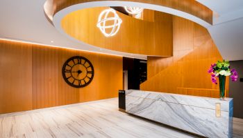 The image shows a modern lobby with a marble reception desk, wooden wall panels, a large wall clock, and decorative lighting fixtures.