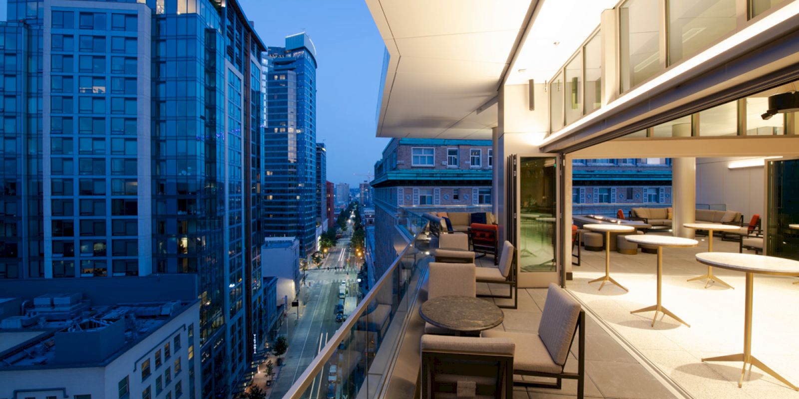 A modern urban rooftop terrace with seating overlooks a city street and high-rise buildings during twilight, providing a scenic view.