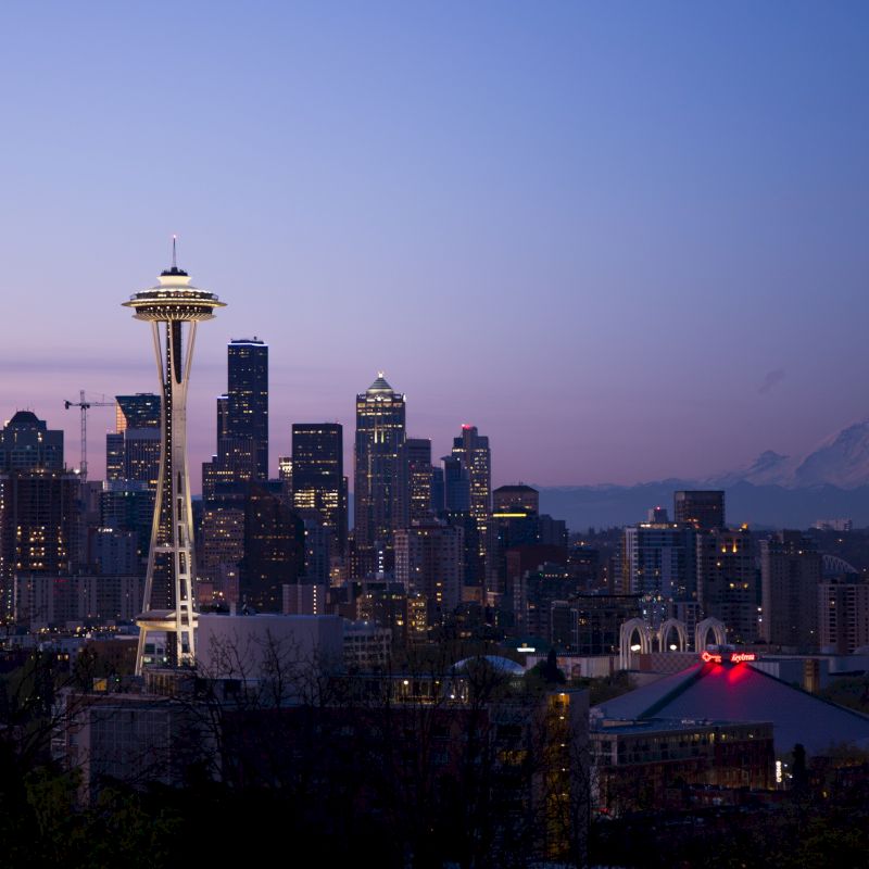 A city skyline at dusk with a prominent tower and mountain in the background, likely Seattle with Mount Rainier visible.