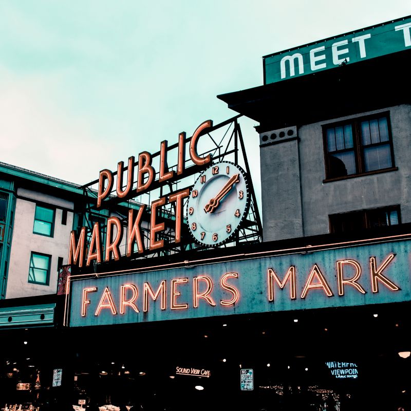 The image shows the iconic neon signs of a public market, with "Farmers Market" and a clock prominently displayed against city buildings.