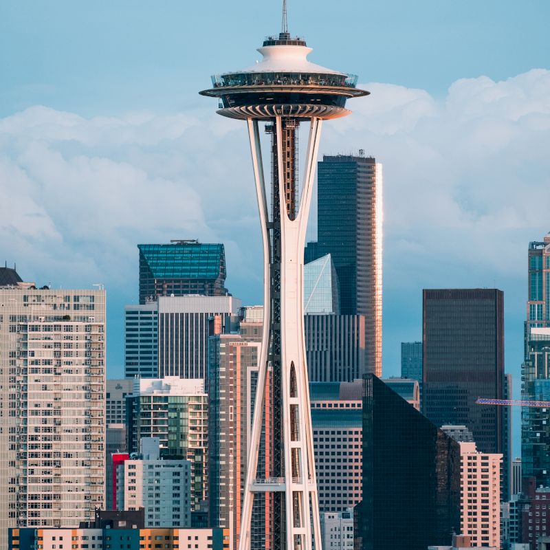 The image shows the Space Needle tower set against a backdrop of Seattle's skyline with several skyscrapers and a cloudy sky.