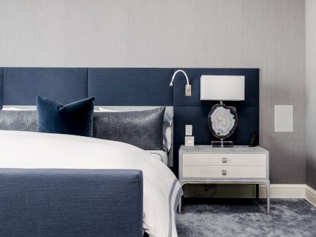 A modern bedroom with a blue upholstered headboard, white nightstand, lamp, and decorative items, all set on a plush carpeted floor.