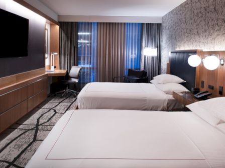 The image shows a modern hotel room with two beds, a flat-screen TV, a desk, lamps, and a large window with curtains.