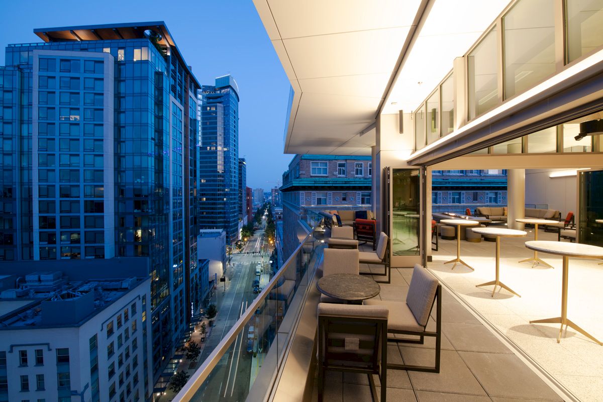 A modern rooftop terrace with seating overlooks a city street surrounded by tall buildings at dusk.