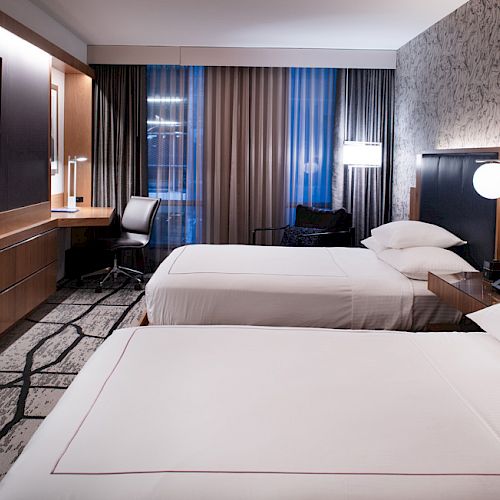 The image shows a modern hotel room with two beds, a wall-mounted TV, a desk, and lamps, featuring a stylish decor and carpeting.