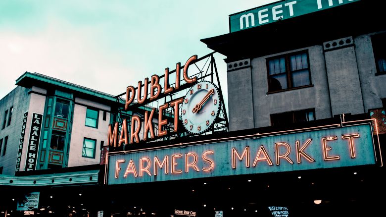 The image shows a public market with a neon sign reading "Farmers Market" and a clock, suggesting a bustling urban setting.
