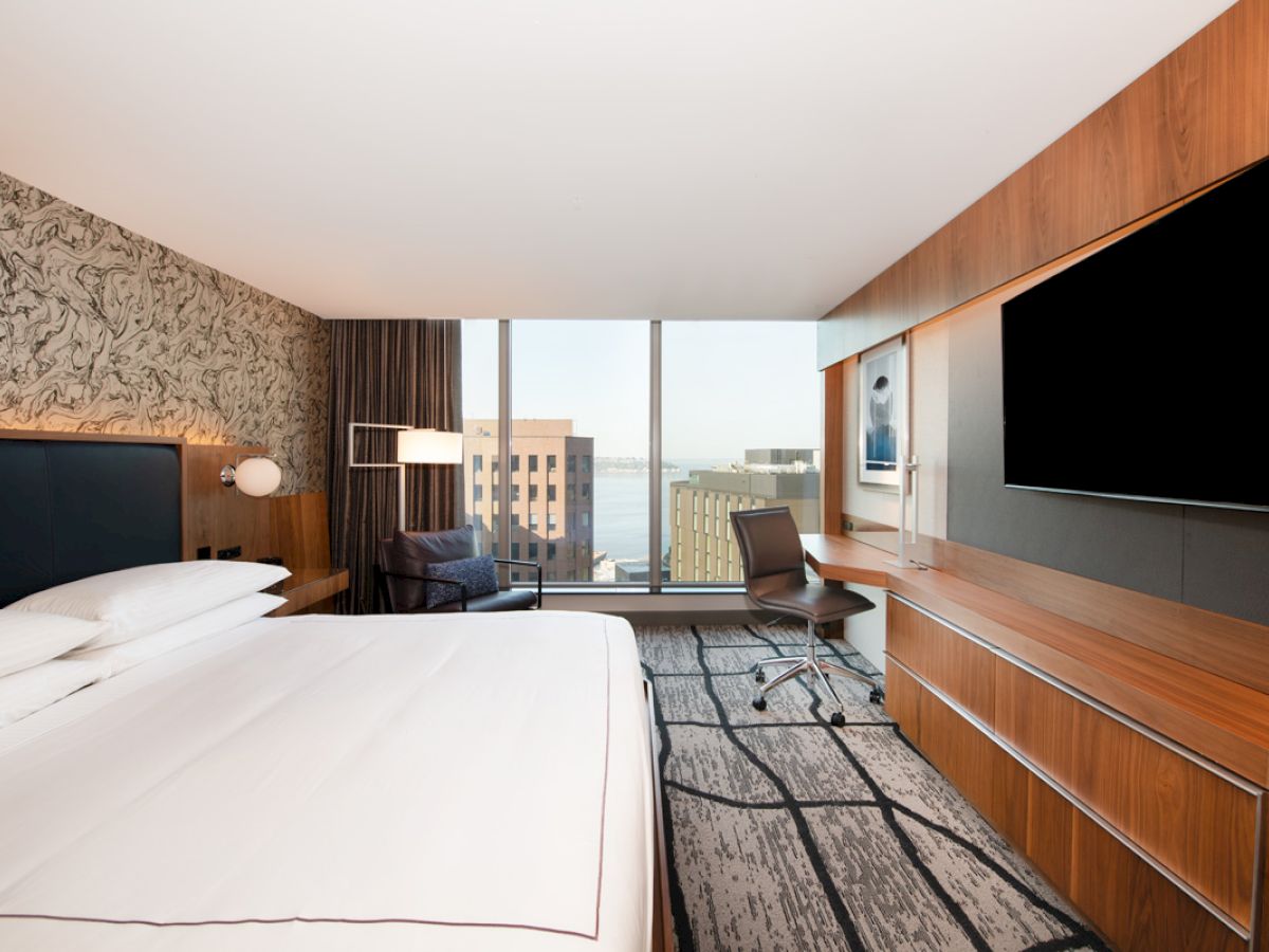 A modern hotel room with a large bed, wall-mounted TV, desk, and a window offering a city view.