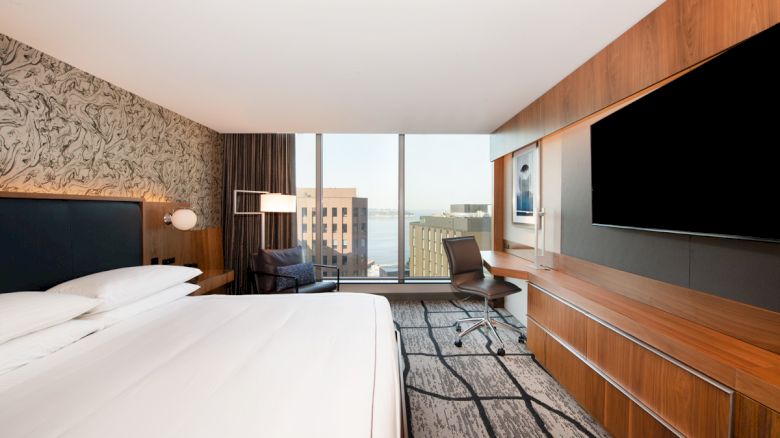 A modern hotel room with a large bed, wall-mounted TV, desk, and a window offering a city view.