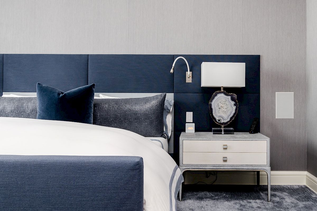 A modern bedroom with a blue and white color scheme features a bed, nightstand with a lamp, and textured wall accents.