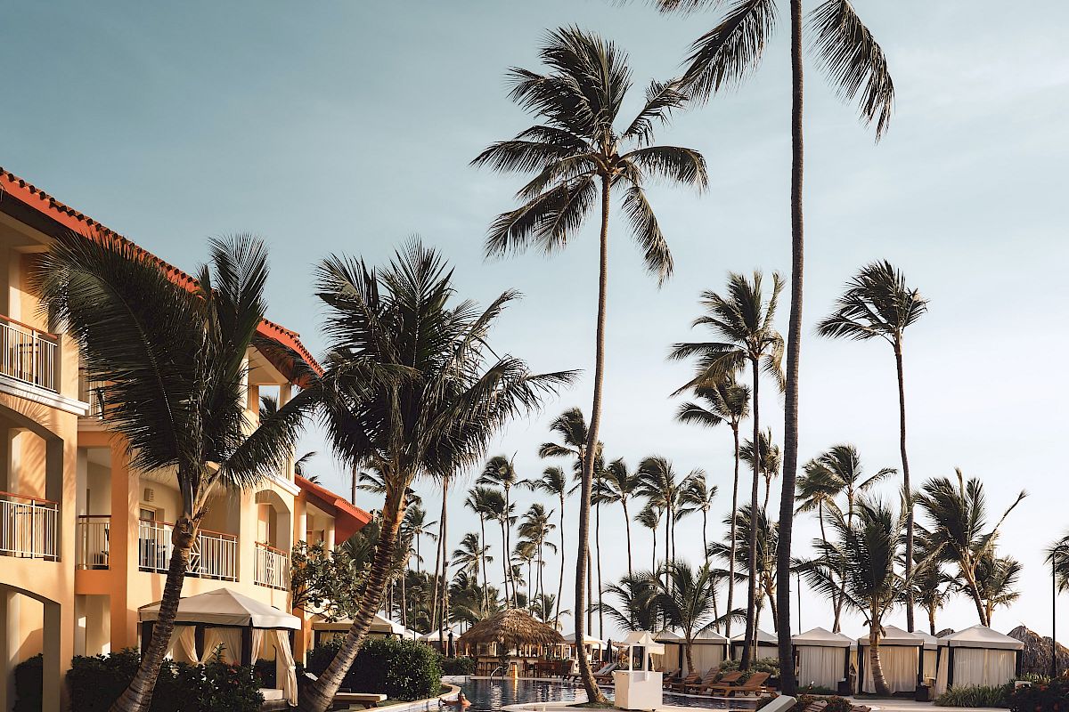 A tropical resort scene with a pool, lounge chairs, palm trees, and a building with red-tiled roofs under a clear sky.