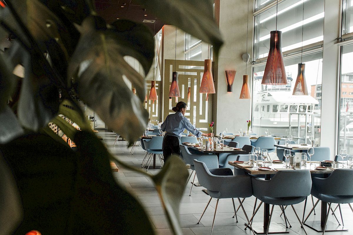 A person sets tables in a modern restaurant with stylish lighting and large windows, partially hidden by plant leaves.