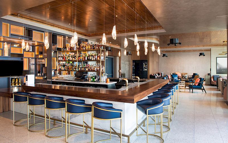 A modern bar with high stools, pendant lights, and stocked shelves in a stylish lounge setting.